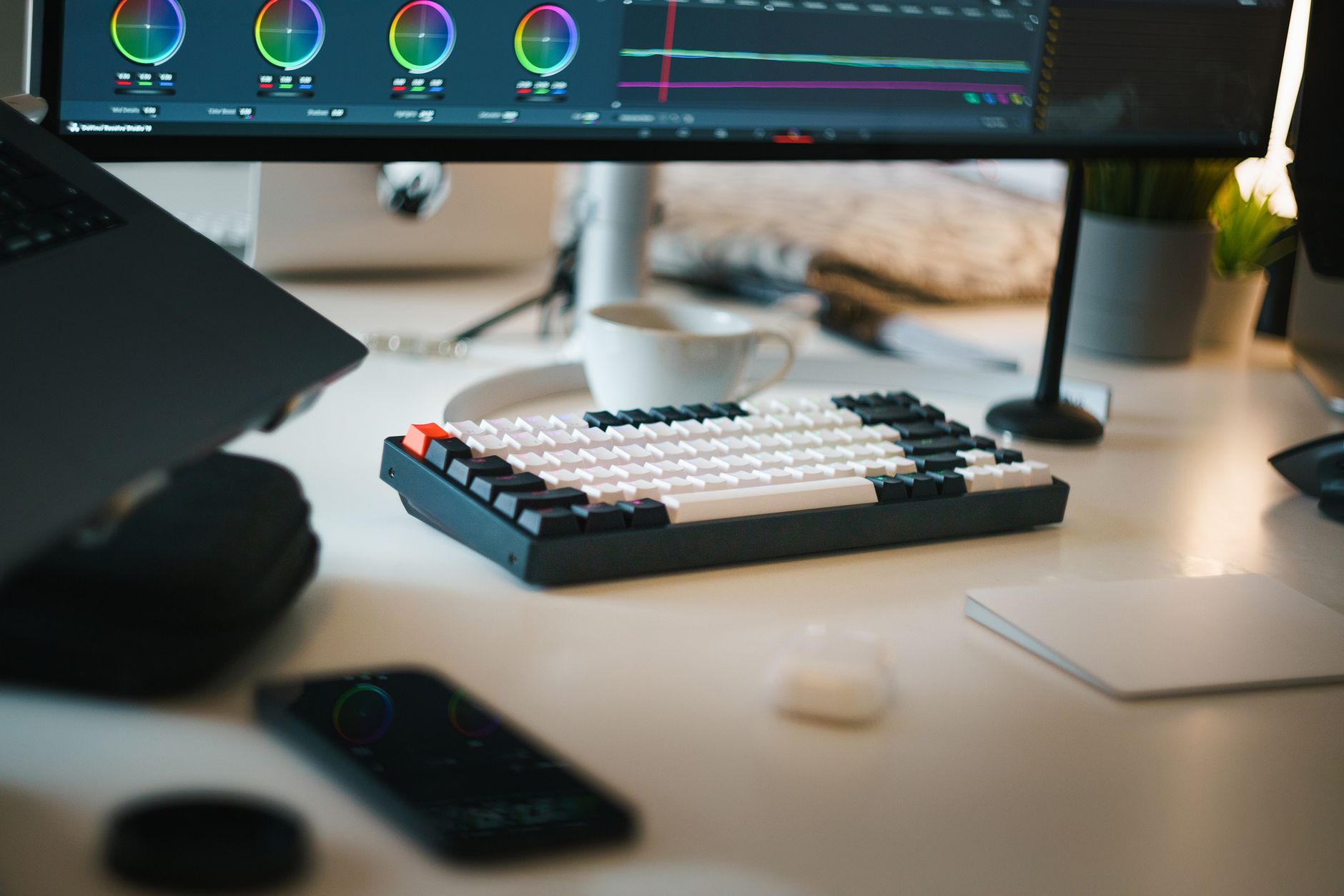 Poste de montage vidéo : écran avec roues colorimétriques, clavier, tasse et smartphone posés sur un bureau clair.