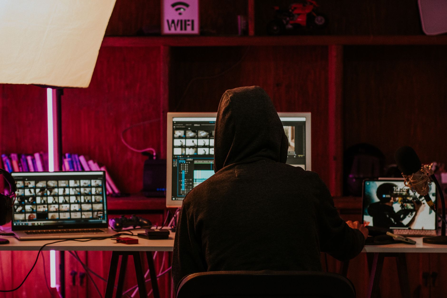 Personne à capuche, de dos, assise devant plusieurs écrans de montage vidéo dans une pièce éclairée en rose.
