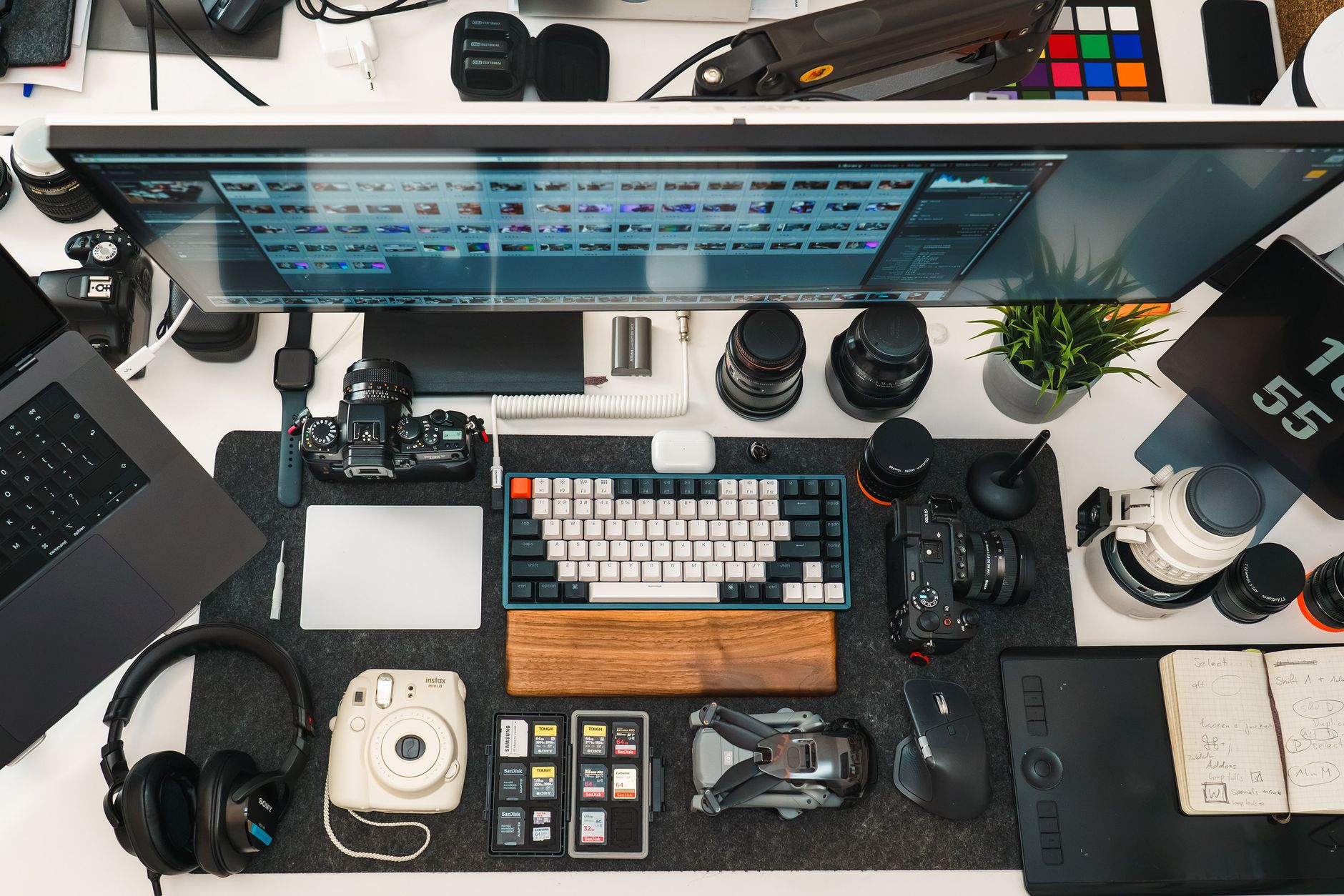 Bureau de créateur vu du dessus : écran de retouche, appareils photo, objectifs, clavier et carnet de notes.